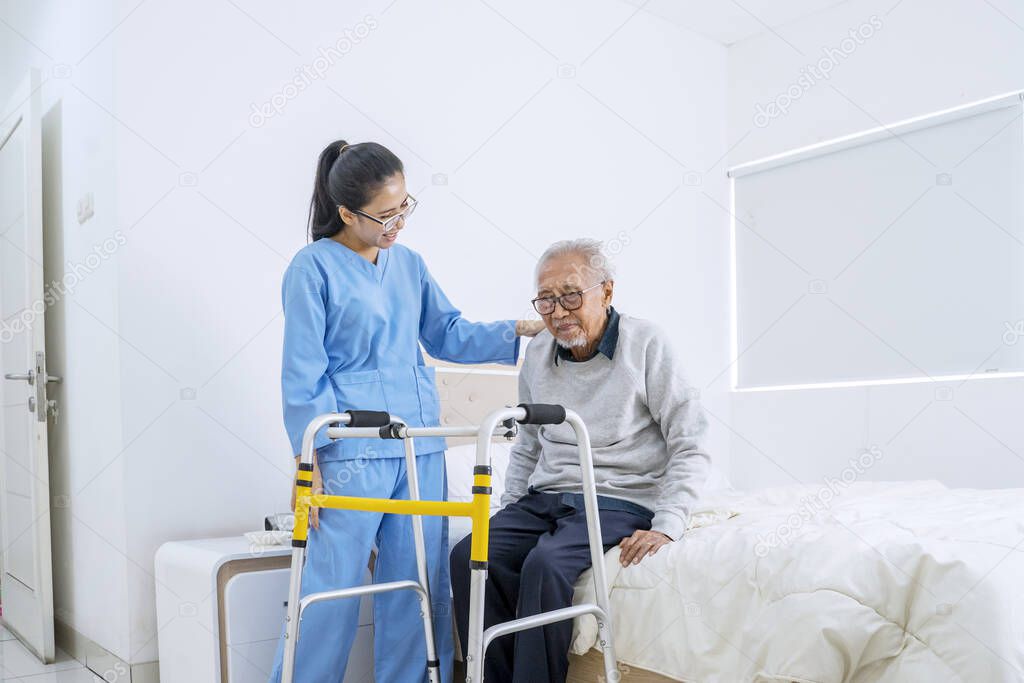 Portrait of senior man sitting on the bed while young nurse helping him with walker equipment