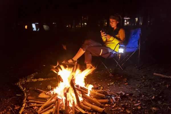 Attractive young woman using cellphone while sitting near the campfire during camping at night on summer time