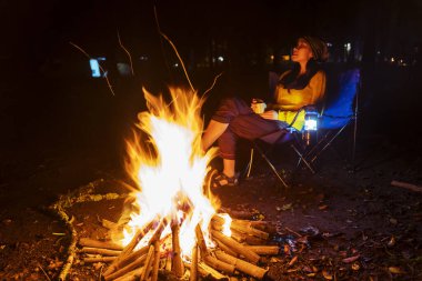Kamp ateşinin yanında dinlenen genç bir kadın yaz gecelerinde kamp yaparken sıcak içkinin tadını çıkarıyor.