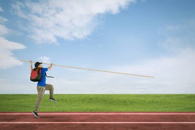 Erkek öğrenci sırıkla atlarken sahada sırt çantası taşıyor.