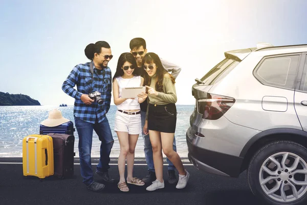 Portrait of cheerful tourists looking at digital tablet while standing behind the car