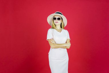 Güzel bir kadının portresi, ellerini şapkalı, güneş gözlüklü, kırmızı arka planlı bir şekilde katlarken kendinden emin görünüyor. 