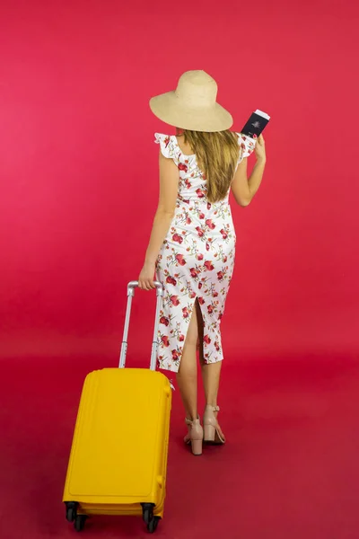 赤を背景にスタジオを歩いている間にパスポートとスーツケースを持っている若い女性のビューの後ろ — ストック写真
