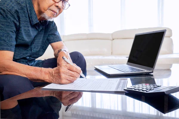 Old Asian man sitting on his sofa while filling and writing the insurance paper form with a pen on the reflecting table, besides calculator and laptop in the living room