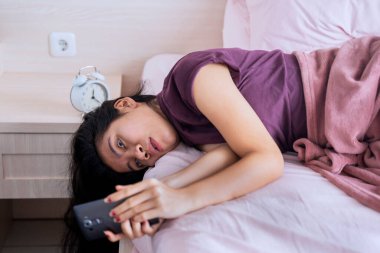 Sıkılırken kendini izole eden ve cep telefonunu yatakta kullanan genç bir kadının portresi.