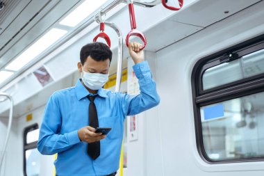 JAKARTA - Endonezya. 12 Mayıs 2020: Genç işadamı koronavirüsü önlemek için maske takıyor cep telefonu kullanıyor ve banliyö treninin içinde duruyor.