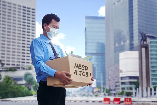 JAKARTA - Indonesia. May 12, 2020: Young businessman holding a cardboard box with text of need work while standing on the sidewalk. Shot with blurred skyscrapers background