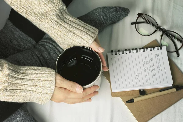 Kobieta w przytulnym sweter, trzymając kubek gorące napoje, pracy i czytając książkę w leniwy dzień, zimowy poranek w domu w łóżku, koncepcja ciepłe uczucie. — Zdjęcie stockowe