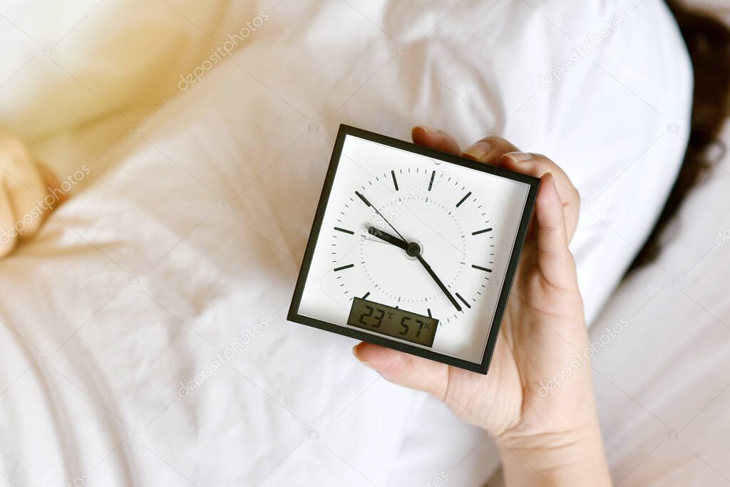 Sleepy girl's hand showing alarm clock and waking up late in the morning, Woman sleep in and laying down in bed hiding under the blanket, Sleep disorder and not enough rest problem.