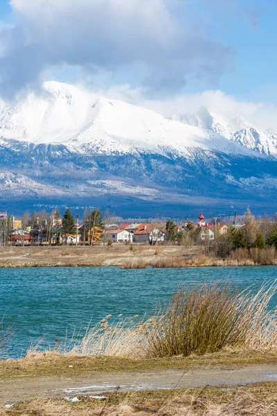 Eslovaquia: Big Tatras (Vysoke Tatry) en primavera. Grandes montañas con nieve y nubes. El lago en primer plano. Pequeño pueblo cerca de la montaña y la colina . — Foto de Stock