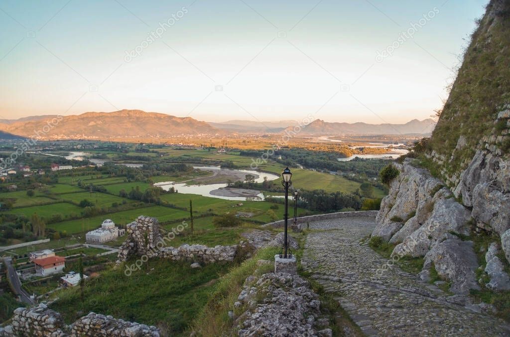 Scenic evening view from the Rozafa Castle. Steep hillside cobblestone street. Old fashioned streetlights. Valley of Drin River and overlooking Albanian Alps in the background. Nature and travel. Albania, Shkoder (Shkodra)