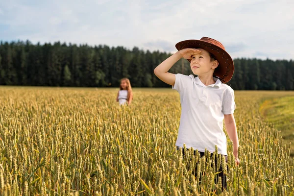 En pojke som ser upp till himlen vid fältet vete i en cowboyhatt och en flicka på en avlägsen bakgrund — Stockfoto
