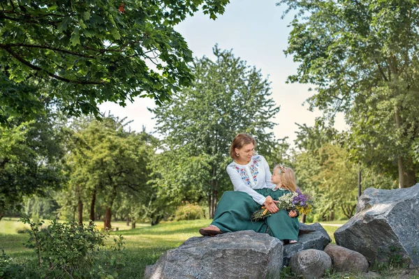 Сімейна мама з дочкою в вінтажному ретро стилі лляні сукні, що сидять на кам'яній скелі в парковому лісі з букетом квітів, дивлячись на очі і посміхаючись — стокове фото