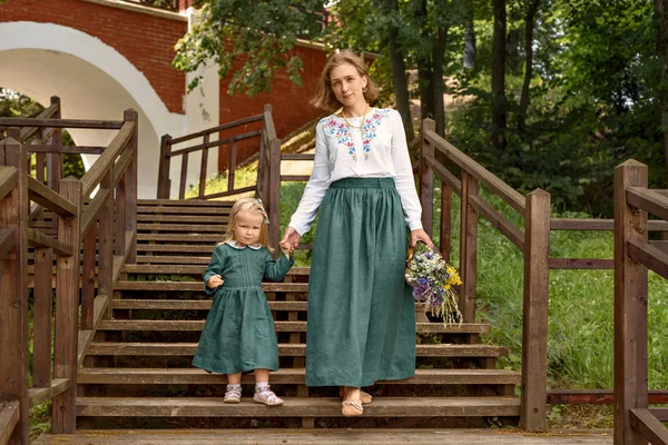 Madre de familia con hija en vestidos de lino de estilo retro vintage con ramo de flores caminando escaleras de madera amanecer en un jardín del parque — Foto de Stock