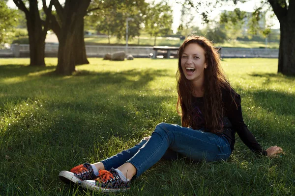 Молодая девушка с рыжими волосами в парке сидит на траве и широко смеется — стоковое фото