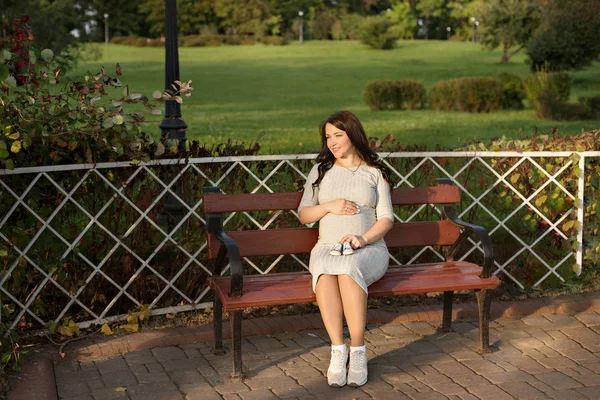 Молодая беременная женщина сидит на скамейке в парке и держит кроссовки для новорожденного — стоковое фото