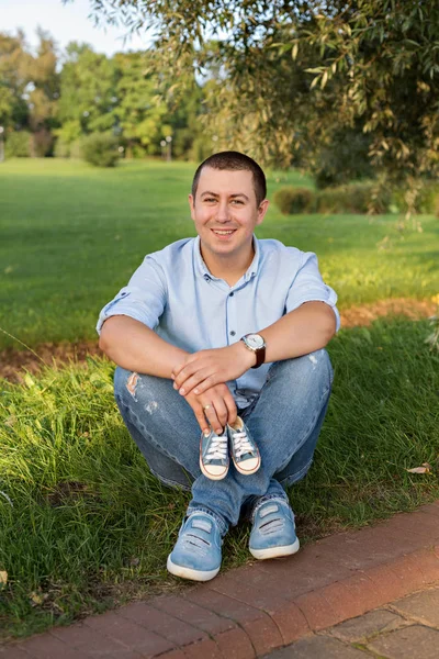 Young man sitting on grass in park and holds sneakers for newborn — Stock Photo, Image