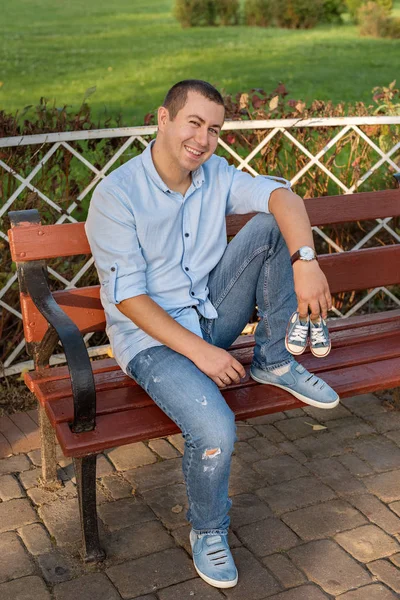 Young man sitting on bench in park and holds sneakers for newborn — Stock Photo, Image