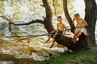 naughty boy and girl sitting on a branch over water fishing, laughing, having fun talking clipart