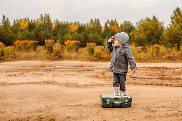 Хлопчик в трусиках і сіре пальто стоїть на старій валізі посеред піщаної сільської дороги, піднімаючи руку на чоло — стокове фото
