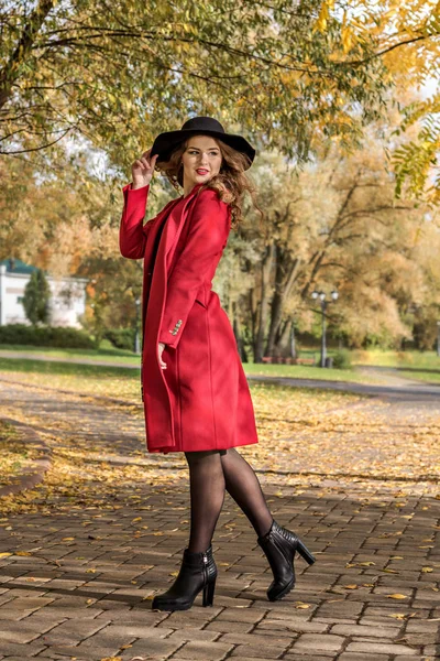 Niña con un abrigo rojo y sombrero se encuentra en un parque de otoño — Foto de Stock