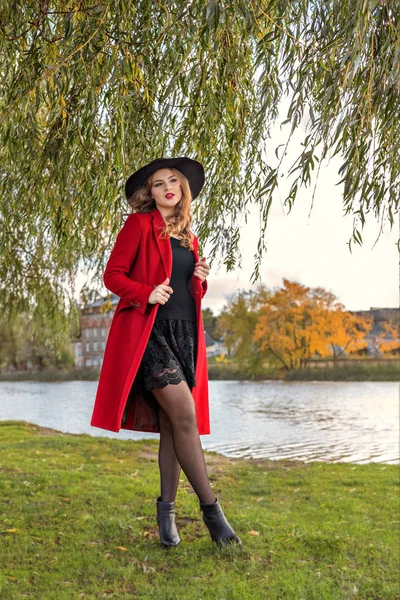 Una niña con un abrigo rojo y sombrero se encuentra debajo de las ramas de un sauce en la orilla del río del lago — Foto de Stock