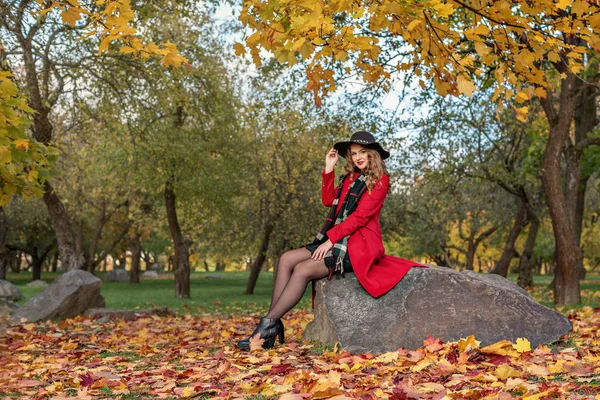 Niña se sienta en una roca en un parque de otoño con un abrigo rojo y sombrero negro en la cabeza — Foto de Stock