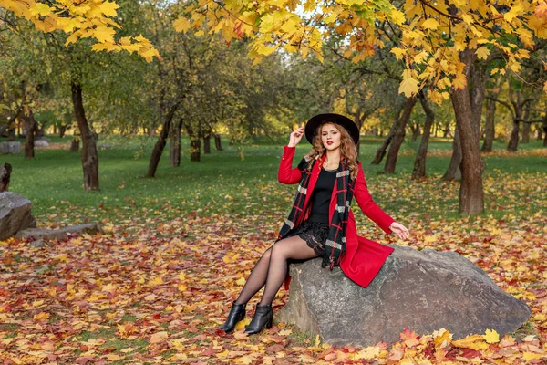 В осеннем парке девушка сидит на камне в красном пальто в черной шляпе и шарфе — стоковое фото