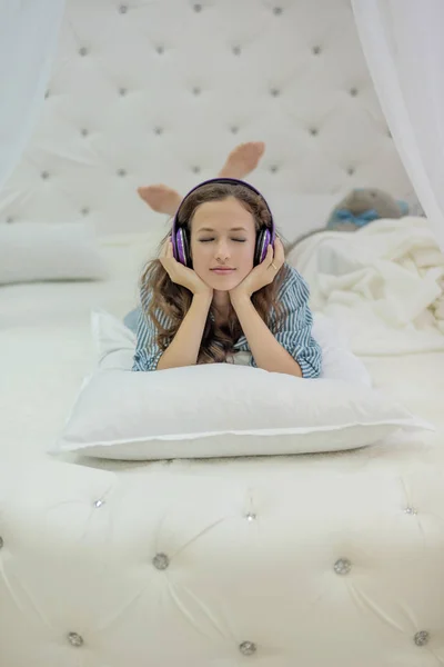 Girl listening to headphones lying on a white bed with bunny ears made with feet — Stock Photo, Image