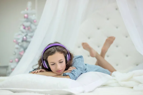 girl lies on a pillow on a white round bed and listens to music on headphones in the bedroom with a new year tree on Christmas Eve