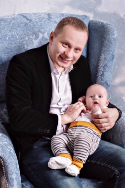 Genç baba, yaşam tarzı bir takım elbiseyle bir sandalyeye oturur ve kucağında bir oğlan çocuğu taşır.