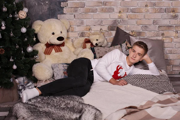 Ένας έφηβος ξαπλώνει σε ένα άνετο κρεβάτι κοντά σε ένα χριστουγεννιάτικο δέντρο με μια μεγάλη αρκούδα. — Φωτογραφία Αρχείου