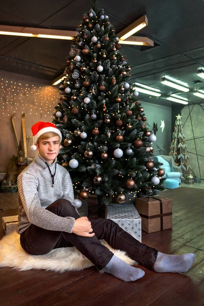 Sorrindo adolescente menino em uma camisola de malha cinza jeans e chapéu de Papai Noel está sentado perto da árvore de Ano Novo e luzes — Fotografia de Stock