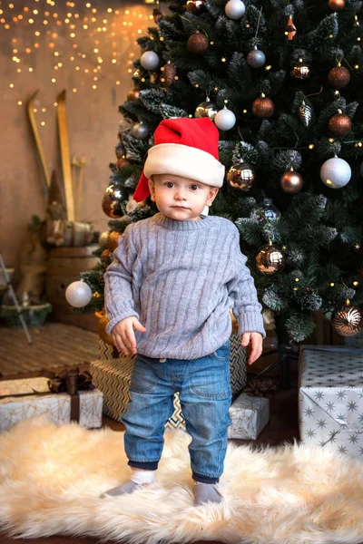 Toddler boy dressed in jeans and a sweater and Santa hat standing near the festively decorated Christmas tree — Stock Photo, Image