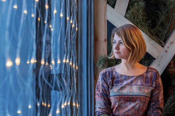 Mujer joven mira por la ventana decorada con una guirnalda de bombillas en la víspera de Año Nuevo — Foto de Stock
