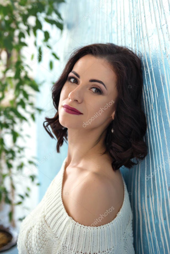 close portrait of a dreaming attractive stylish woman in a white sweater on one shoulder near the blue wall and a green plant on the background