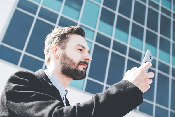 Bearded modern man with wireless headphones in his ears listening to music from a smartphone and walking on outdoors — Stock Photo, Image