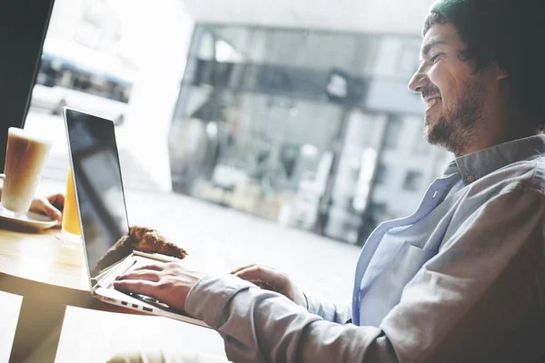Vrolijke en jonge man in casual kleding zitten in een cafe tegenover een groot raam en werken voor een laptop — Stockfoto