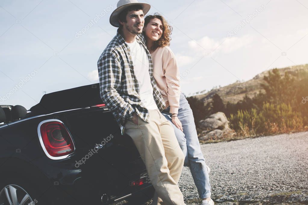 Modern young couple is traveling on a convertible car, they rest and admire the mountain road on a warm sunny day