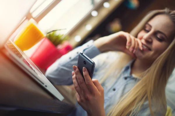 Hermosa mujer freelancer comprueba las noticias en las redes sociales en su teléfono en un acogedor café en una mesa con un ordenador portátil y un vaso de jugo de naranja —  Fotos de Stock