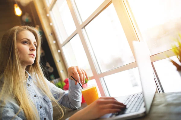 Hermosas mujeres freelancer sueña con algo relajante sentado en un café cerca de una gran ventana en una mesa con un ordenador portátil y un vaso de jugo de naranja — Foto de Stock