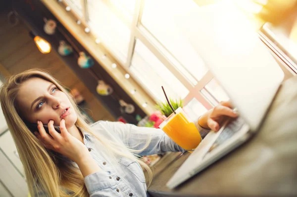 Pensive woman blogger working at a laptop and talking about something on the phone while sitting at a table with a glass of orange juice near a large window in a modern cafe — Stock Photo, Image