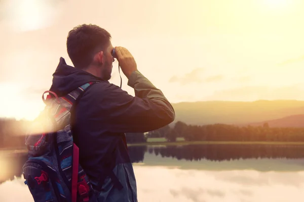 Viajante com uma mochila olhando através de binóculos na montanha e no lago — Fotografia de Stock