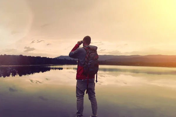 Молодий чоловік з рюкзаком стоїть біля озера і дивиться через бінокль — стокове фото