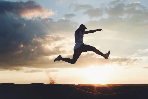 Silhouette of a strong athlete jumping over the hills. Man at sunset in nature is engaged in fitness. Intentional dark colors — Stock Photo, Image