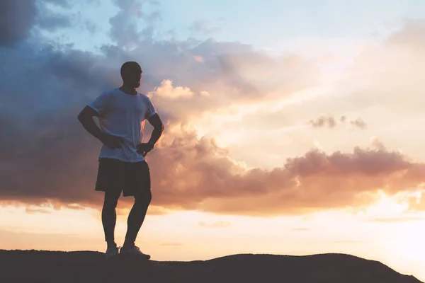 Homem de roupas esportivas está de pé em uma colina após o treinamento ao pôr do sol — Fotografia de Stock