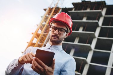 Modern inşaat mühendisi veya mimar inşaat sahasında kask içinde endüstriyel elektronik tablet ile çalışır. Genç bir mühendis ile becerikli alet inşaat binasının arka plan üzerinde yakın çekim portresi