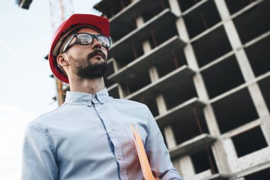 Modern business man in red hard hat holds folder with blueprint for construction industrial factory. Young bearded architect inspects construction of new building clipart