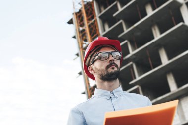 Bina İnşaatı için belgeleri içeren paketi ile kırmızı kask içinde mühendis Oluşturucu. İnşaat sahasında mimar ya da genç başarılı erkek inşaat işçisi