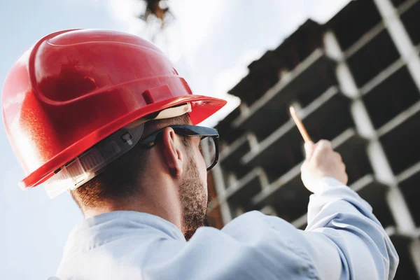 Inspector inspecciona la construcción de rascacielos. Ingeniero masculino o oficial de seguridad en casco rojo en el fondo de la obra — Foto de Stock
